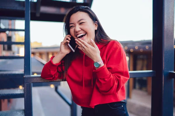 Happy excited female teengaer dressed in trendy red hoody laughing during friendly smartphone conversation via application and roaming connection in public place, cheerful chinese woman using mobile