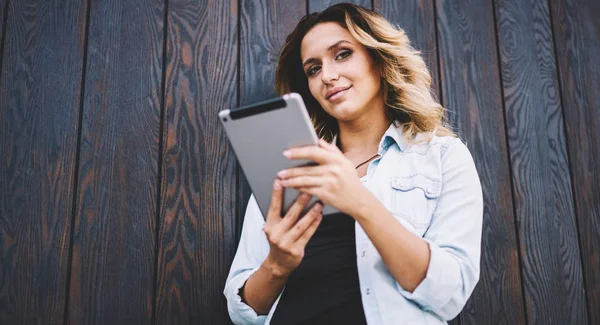 Half length portrait of positive smiling hipster girl with digital tablet in hands looking at camera while posing near copy space area for advertising text, happy female using gadget for communicate