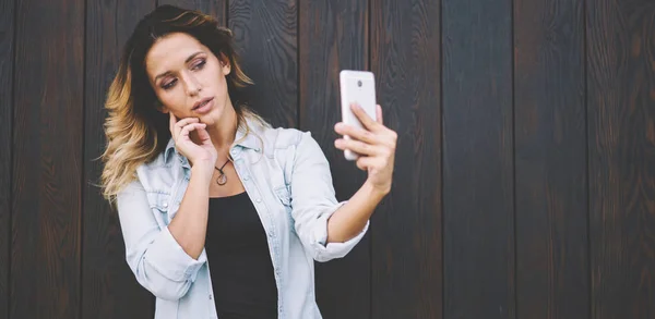 Millennial beautiful female teenager making sexy photos for own blog on internet space, attractive woman using front camera for clicking selfie pictures near copy space area for advertising text