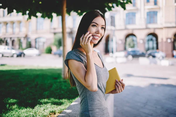 Positive hipster girl happy to hear friendly voice during international telephone conversation outdoors, smiling female teenager with notebook talking with colleague via cellphone on way to university