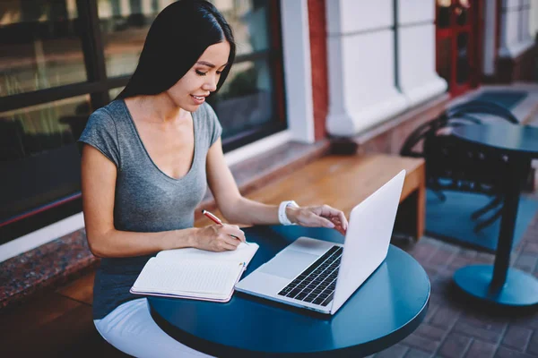 Positive female journalist writing article for magazine in knowledge textbook while sitting at cafeteria table with modern computer, attractive woman student making notes for university course work