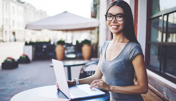 Portrait of happy female freelancer smiling and sitting with laptop computer in street cafeteria during summer day, cheerful woman in optical spectacles enjoying distance job with modern netbook