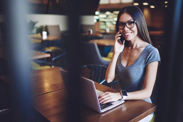 快乐的女人打电话到客户服务咨询关于笔记本电脑的应用 成功的时髦的女孩在眼镜享受时间在共同工作空间 而通过手机沟通 — 图库照片