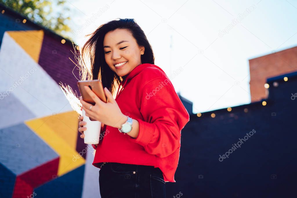 Concept of youth lifestyle and generation, happy cheerful chinese hipster girl reading received sms message and smiling from funny text while enjoying time for coffee beverage on sunny city street