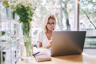 Laptop cihazı ile kafeteryada otururken sohbet için akıllı telefon uygulamasında bildirim güncelleme göz gözlük gülümseyen kadın, bin yıllık hipster kız hücresel iyi WiFi bağlantısı zevk