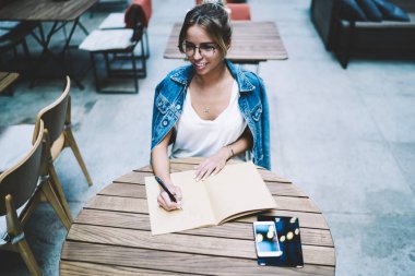Keyifli kadın yazar, kafeterya masasında eğitim için Notebook ile oturan heyecanlı bir fikir ile kompozisyon yaratma, göz giyim çekici gülümseyen hipster kız otodidakt kapalı ücretsiz zaman zevk