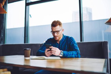 Kafkas hipster adam kahve dükkanında öğrenme mola sırasında modern cep telefonu üzerinden sosyal ağ da haber okuma, genç erkek öğrenci masada ders kitabı ile oturan ve mobil uygulama yükleme