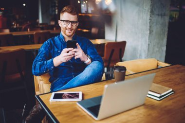 Gözlük pozitif erkek blogger portresi elinde cep telefonu ile oturan ve modern netbook, nesil ve iletişim kavramı üzerinde uzaktan çalışma dan mola sırasında kamera gülümseyen