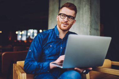 Dizüstü bilgisayarda internet bağlantısı beklerken uzaklara bakarak göz koruması sağlamak için optik gözlük düşünceli hipster adam, coworking alanında netbook ile oturan adam düşünme