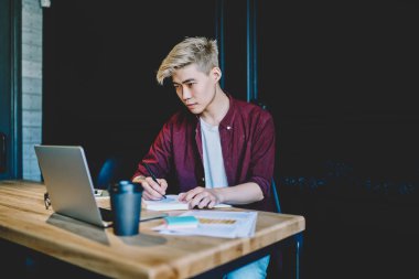 Ciddi Japon hipster adam otomasyon için zaman harcıyor birlikte çalışma alanında bilgi verici webinar izliyor modern dizüstü bilgisayarla ve eğitim için ders kitabına ana notlar yazıyor.