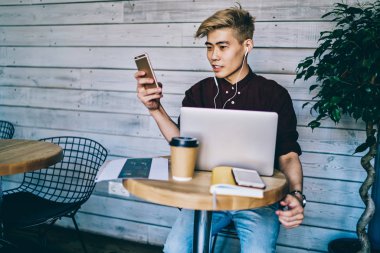 Havalı hipster adam akıllı telefon uygulaması aracılığıyla mesaj aldı. Elektronik kulaklıklar aracılığıyla müzik listesi dinlerken kafeteryada modern dizüstü bilgisayarla oturuyordu.