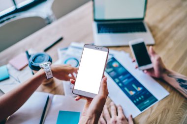 Kağıt grafik, diyagram ve dizüstü bilgisayarlı ahşap masaüstüyle arka planda internet içeriğiniz için boş ekran alanı olan modern akıllı telefonun el ele tutuşunun kırpılmış görüntüsü.