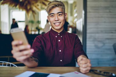 Neşeli gülümseyen Asyalı öğrenci elinde cep telefonuyla masaüstünde oturuyor ve 4G internet bağlantısı kullanıyor, mutlu Japon hipster adam cep telefonuyla çevrimiçi hizmetten geri bildirim bekliyor.