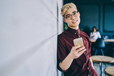 Modern cep telefonunu elinde tutan ve boş zamanlarında kameraya gülümseyen optik gözlük takan binlerce yıllık erkek blog yazarının yarım boy portresi.