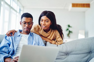 Genç Afro-Amerikalı çift internet sayfasından alışveriş yapıyor dizüstü bilgisayarla evde kalıyor, gülümsüyor siyah tenli hipsterler evde boş zamanlarında netbook 'ta video izliyorlar.
