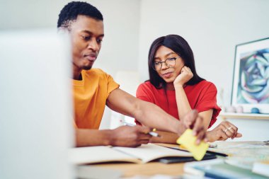 Gülümseyen Afro-Amerikan kadın kocasının evde bağımsız olarak çalışmasını izliyor. Pozitif esmer tenli kadın, masaüstü öğrenme ve not alma konusunda arkadaşlarıyla iş birliği yaparak tatmin oluyor.