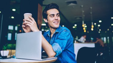 Neşeli hipster adam kulaklık ile olumlu ses kaydı zevk sırasında paket kahve tutan, genç gülümseyen adam müzik dinlerken ve dizüstü bilgisayarda iletişim mola uzağa bakıyor
