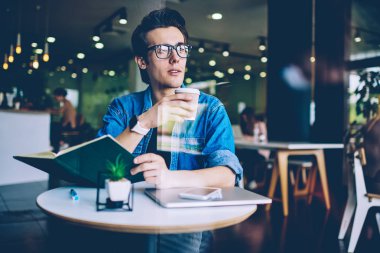 Sınava hazırlanmak için bilgi literatürü kullanarak kapalı mekanda kafeinli içecek keyfi gözlük genç adam Pondering, düşünceli hipster adam rahat paket kahve tutarak giyinmiş
