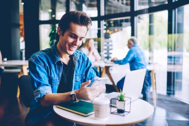 Kafeterya ücretsiz kablosuz bağlantı kullanarak mobil uygulama üzerinden komedi gösterisi online izlerken Neşeli hipster adam, sosyal ağ için app yeni bildirim iyi duygu gülümseyen erkek blogger