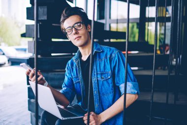 Akıllı telefon aletini elinde tutan ve mobil etkin noktadan dizüstü bilgisayarda 4g internete bağlanmaya çalışırken kameraya bakan optik gözlüklü genç yakışıklı hipster adam portresi