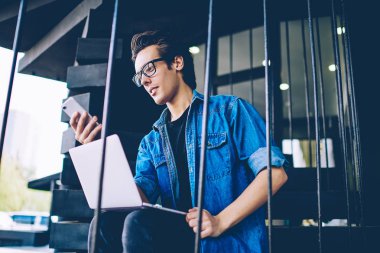 E öğrenme için modern dizüstü bilgisayar ile açık merdivenlerde otururken cep telefonu hotspot bağlantısı hakkında göz koruması kontrol bildirimi sağlamak için gözlük genç hipster adam