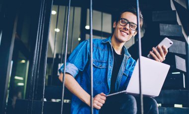 Akıllı telefon cihazı tutarken ve 4g internet bağlantısı hakkında danışmanlık için online müşteri hizmetinden cevap beklerken gözlük neşeli hipster adam Portresi kamera bakıyor