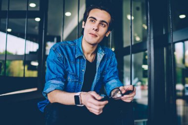 Rahat kıyafet giymiş hipster adam elinde optik gözlük tutan ve düşünceli serbest zaman sırasında uzağa bakarak, dalgın erkek öğrenci arkadaş beklerken üniversite sınavları hakkında düşünme