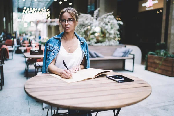 Pensive woman pondering on new article for blog about restaurants while sitting at cafeteria with cozy interior, thoughtful hipster girl in optical eyeglasses looking away and thinking about project