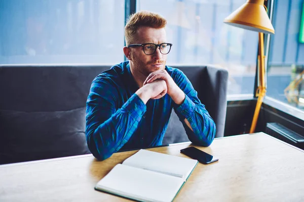 Thoughtful hipster guy in optical eyewear for vision correction pondering on ideo for university project, contemplative man in stylish eyeglasses sitting at desktop with textbook for making notes