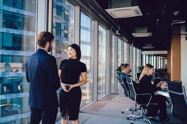 Successful smiling woman talking to male employee while standing in office interior near skyscraper window, corporate positive colleagues enjoying live communication indoors. Two business people