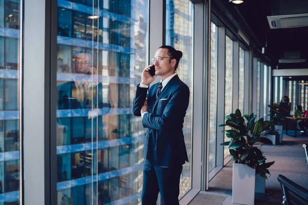 Caucasian male financial director of company making important smartphone call while standing near office window, half length of mature businessman in suit communicating via mobile phone