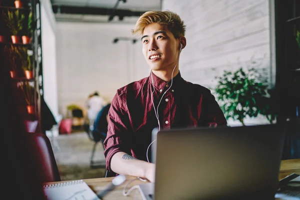 Prosperous japanese hipster guy with stylish blonde hairstyle listening motivation audio books via electronic earphones during working remotely with laptop computer in modern coworking space