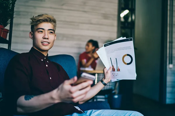 Chinese blonde hipster guy dressed in casual wear pondering on received email from business partner with new information about startup project while working remotely and analyzing paper graphic