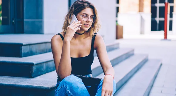 Positive female student communicate with friend after university via smartphone gadget on urban setting, attractive hipster girl calling to friend using mobile application and 4g internet in roaming