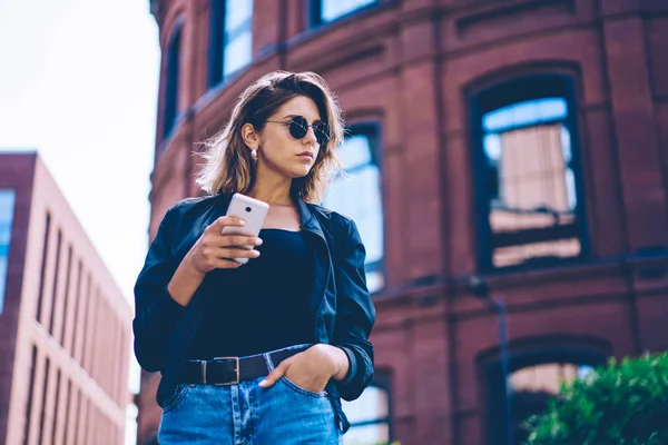 Confident woman in sunglasses looking away while holding smartphone gadget and waiting for call from taxi service, trendy female tourist spending free time for travelling and using telephone