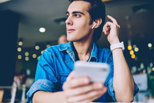 Thoughtful hipster guy looking away and feeling pondering on audio record about motivation, young man holding smartphone gadget in hands while enjoying time for listening music via earphones
