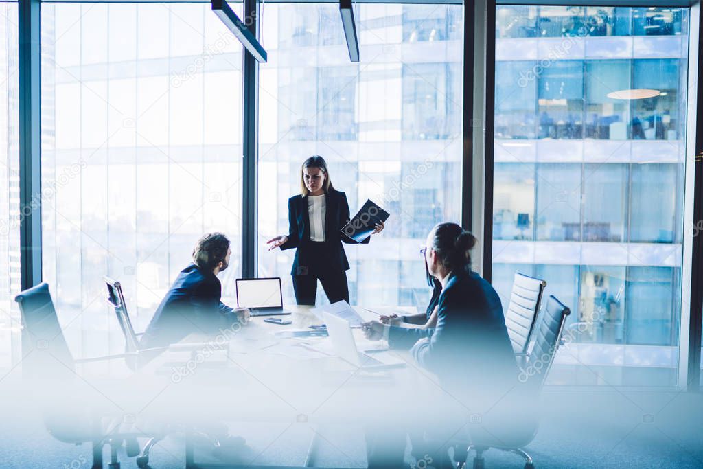 Puzzled female director in elegant suit communicating with employees during working process in conference room standing near meeting table with modern technology, laptop with mock up for website