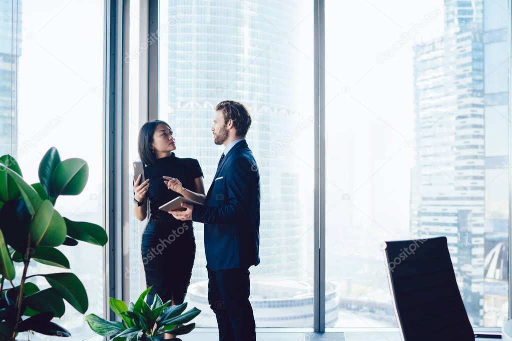 Successful diverse male and female employees in formal wear discussing financial ideas while standing near office window of skyscraper building. Corporate man and woman colleagues talking about work