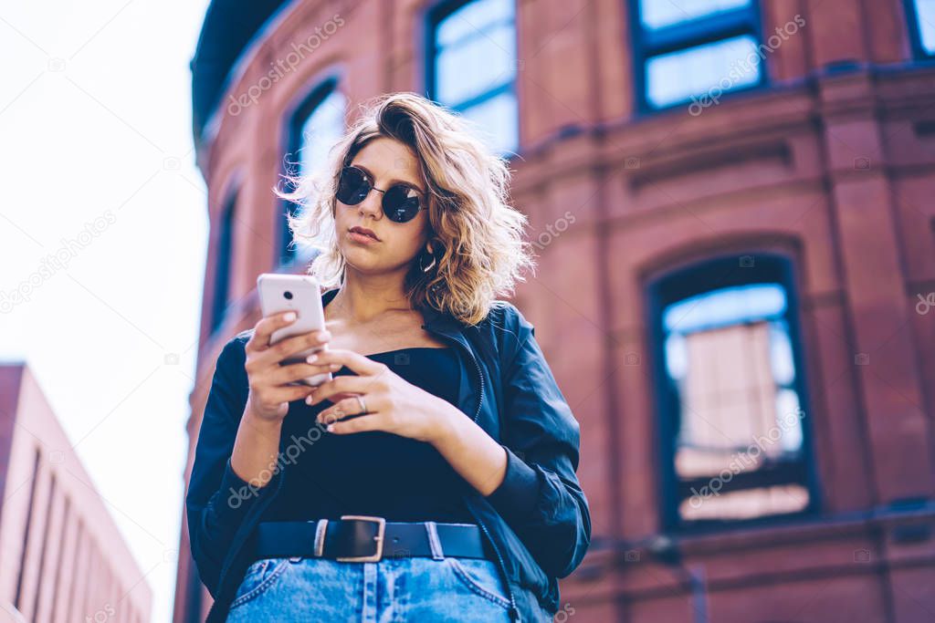 Confident travel blogger publishing publication with content article about tourism using 4g wireless internet on cellphone, attractive hipster girl in sunglasses installing application on telephone