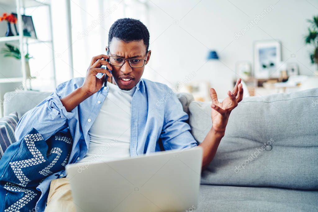 Angry African American hipster guy arguing with operator disappointed with bad wireless connection on netbook for remote job at home, stressed dark skinned man calling via mobile phone having trouble