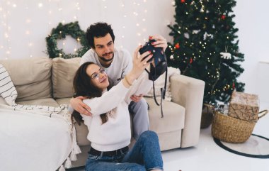 Gülümseyen beyaz kız arkadaş ve erkek arkadaş, kış tatillerinde fotoğraf çekmek için poz veriyor, yeni yılı kutluyor, romantik çift evde noel arifesinde fotoğraf çekmek için kamerayı kullanıyor.