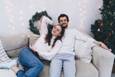 Gülümseyen beyaz erkek ve kadın çift kış tatillerinde şenlikli bir dairede kanepede dinleniyor. Yeni yılı kutlayan kameraya bakarak pozitif evlilik yapıyorlar.