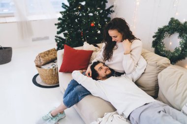 Gündelik giysiler içinde genç bir kadın, erkek arkadaşıyla boş vakitlerini parlak oturma odalarında dinlenerek geçiriyor. Pastel renklerle süslenmiş, Noel ışıkları ve ağaçlarla süslenmiş.