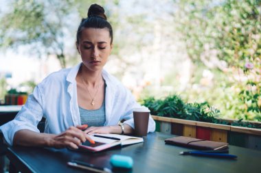 Düşünceli bir kadın, günlük giysiler içinde açık kafedeki ahşap masada oturuyor ve hafta sonu dinlenirken akıllı telefonuyla mesajlaşıyor.