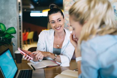 Siyah saçlı neşeli bir kadın internet işi sırasında bayan meslektaşıyla birlikte otururken cep telefonundan bilgi topluyor.