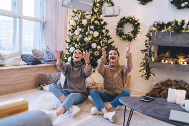 Modern bir dairede Noel hediyeleriyle oynarken pencerenin yanında oturan gülümseyen bayan arkadaşlar.