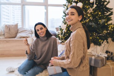 Sıcak süveter giymiş neşeli kadınlar Noel ağacının yanında oturmuş Noel tatilinin tadını çıkarıyorlar.