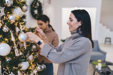 Sıcak giysiler içinde gülümseyen bir kadın arkadaşlarıyla Noel ağacı süslerken iç dekorasyonlarla dolu rahat bir odada bulunan çelenkleri aydınlatan bir çelenk.