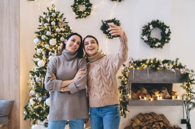 Sıcak giysiler içinde mutlu bayan arkadaşlar akıllı telefondan selfie çekerken aydınlık odada süslenmiş Noel ağacına karşı durup kış tatilini kutlamaya hazırlanıyorlar.