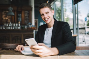 Modern kafede bir fincan kahve içip kameraya bakarken şık kıyafetli, dişlek gülüşlü genç adam akıllı telefon kullanıyor.